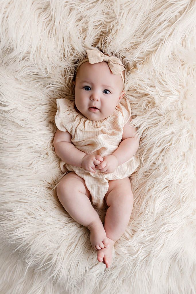 Adorable baby girl lying on fluffy cream rug wearing beige ruffle romper and bow headband.
