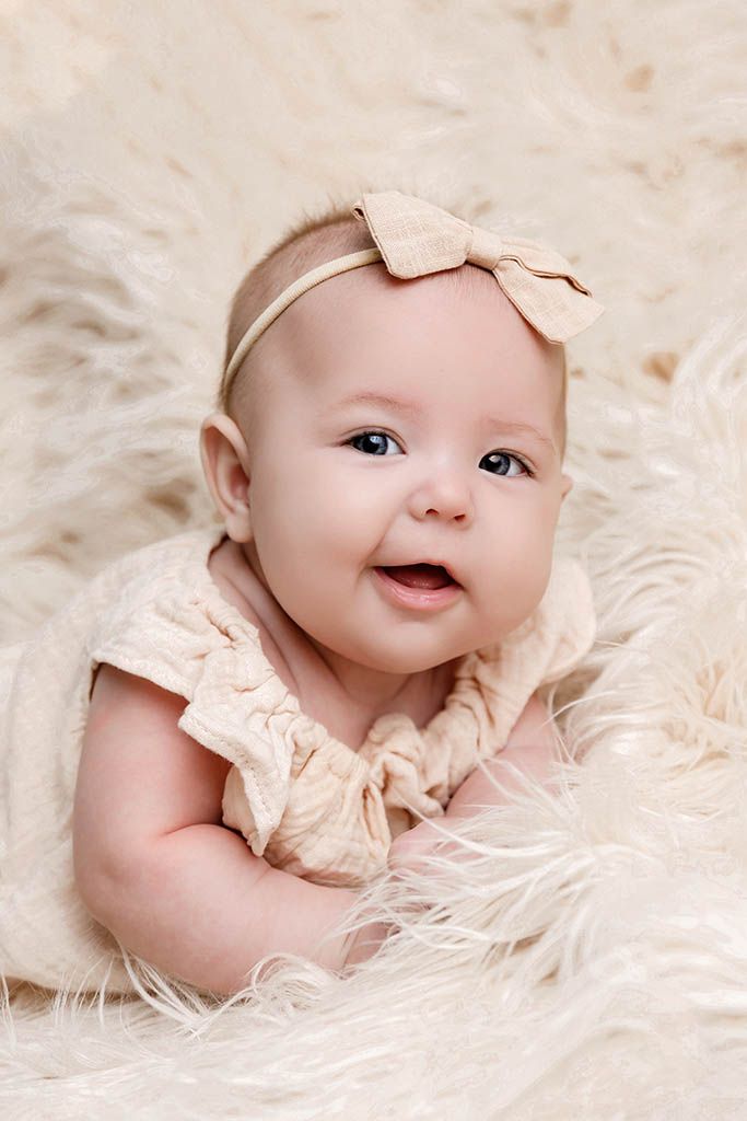 Smiling baby girl in cream outfit and bow headband lying on fluffy white fur blanket.