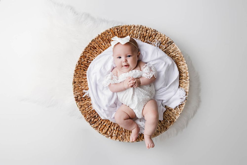 Cute baby girl in white lace outfit lying in wicker basket with bow headband.