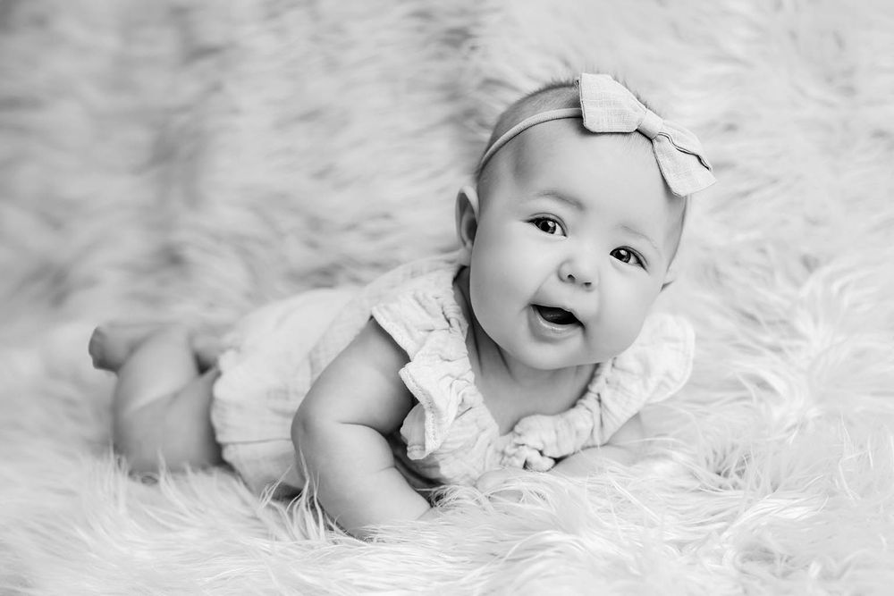 Black and white photo of a smiling baby girl in a ruffled onesie and bow headband lying on a fluffy white rug.