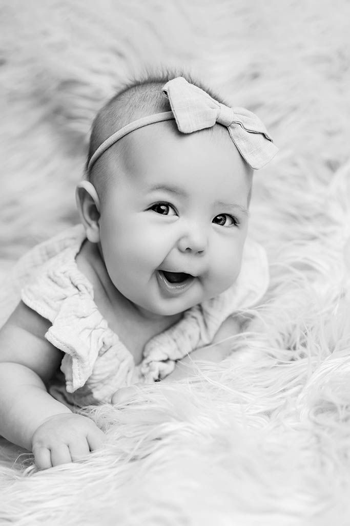 Black and white photo of a smiling baby girl wearing a bow headband, lying on a fluffy white fur blanket.