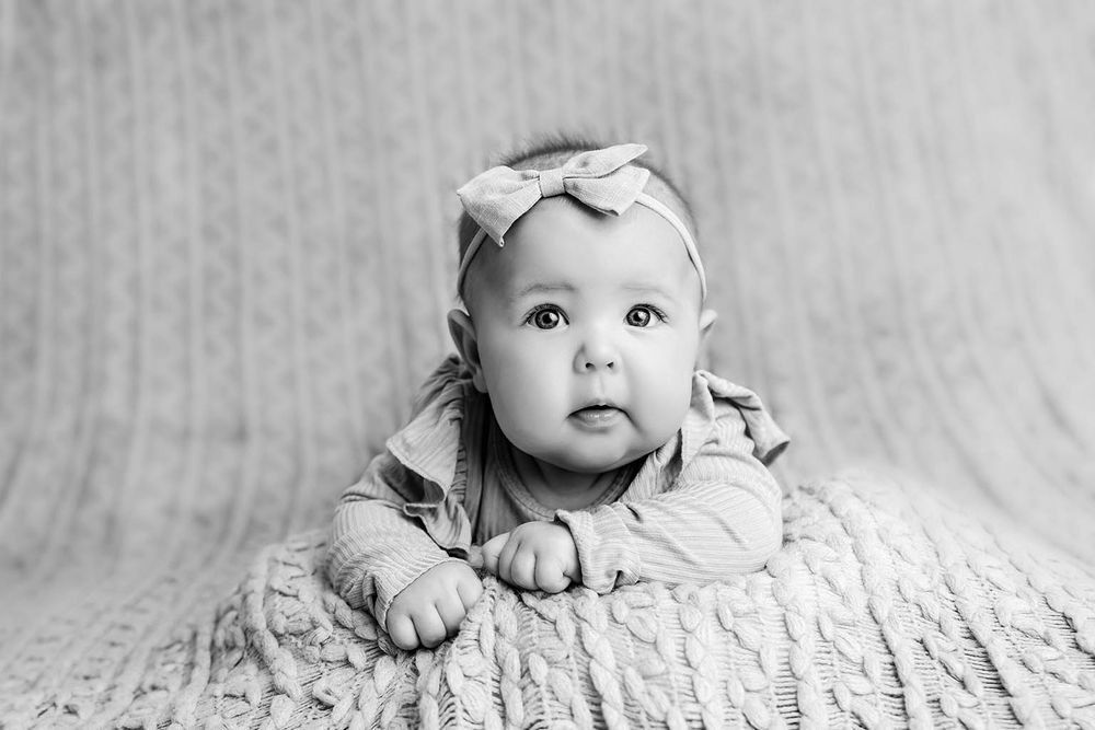 Black and white photo of an adorable baby girl wearing a bow headband, lying on a knit blanket.