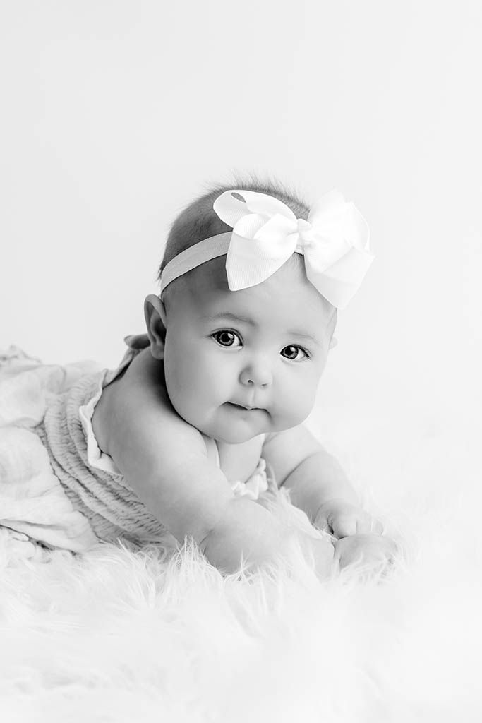 Black and white portrait of an adorable baby girl wearing a white bow headband, lying on a fluffy white blanket.