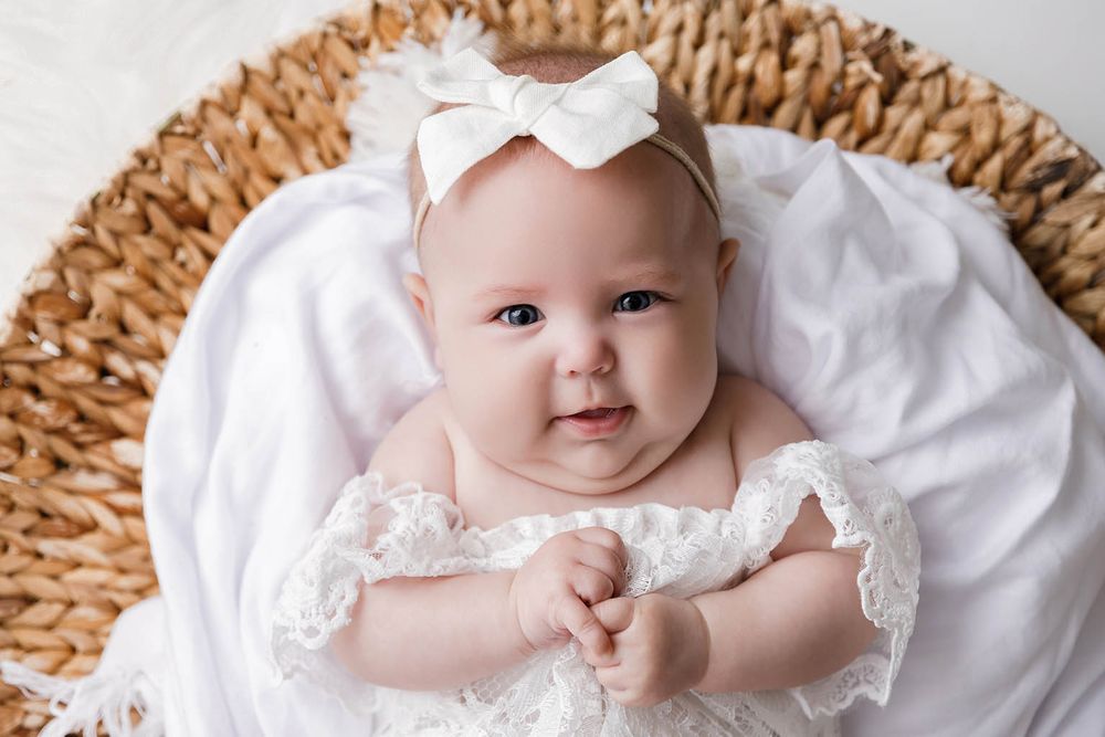 Adorable baby girl in white lace dress and bow headband lying in wicker basket with white blanket.
