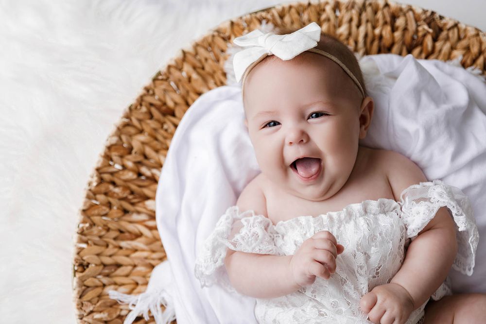 Smiling baby girl in white lace dress and bow headband lying in wicker basket.