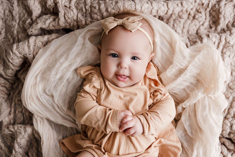 Sweet baby girl in tan outfit with matching bow headband smiling on beige blanket.