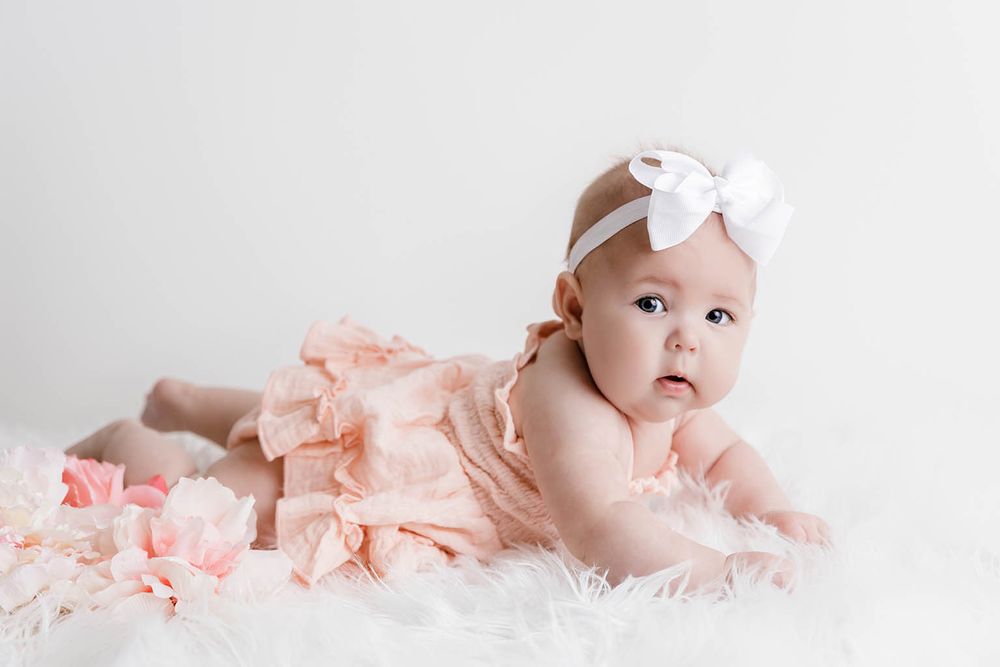 Cute baby girl in pink ruffle dress and white bow headband lying on fur blanket.