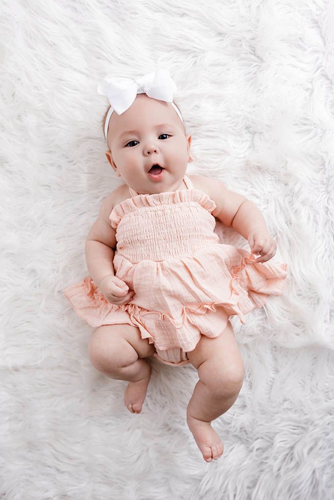 Adorable baby girl in pink ruffle romper with white bow headband lying on fluffy white rug.