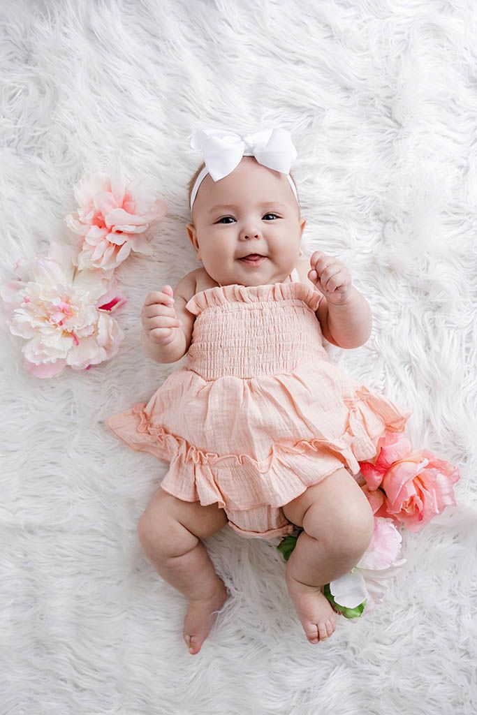 Happy baby girl in pink ruffle romper with white bow headband lying on white fur with flowers.