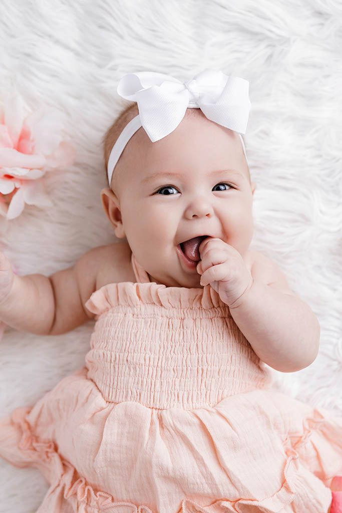 Happy laughing baby girl in peach smocked romper with white bow headband on fluffy white rug.