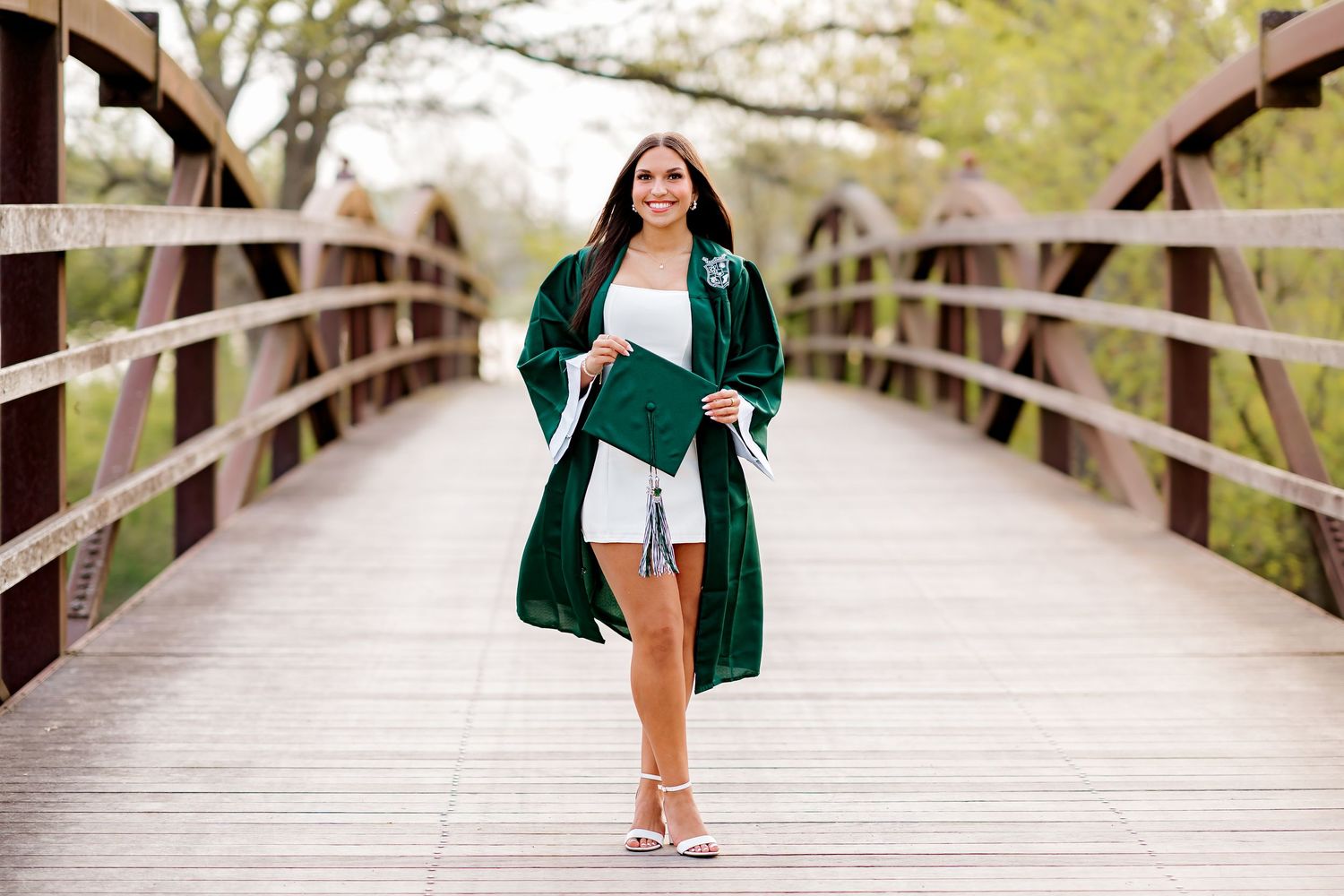 Young woman in green graduation gown and cap poses on wooden bridge during outdoor senior portrait session.