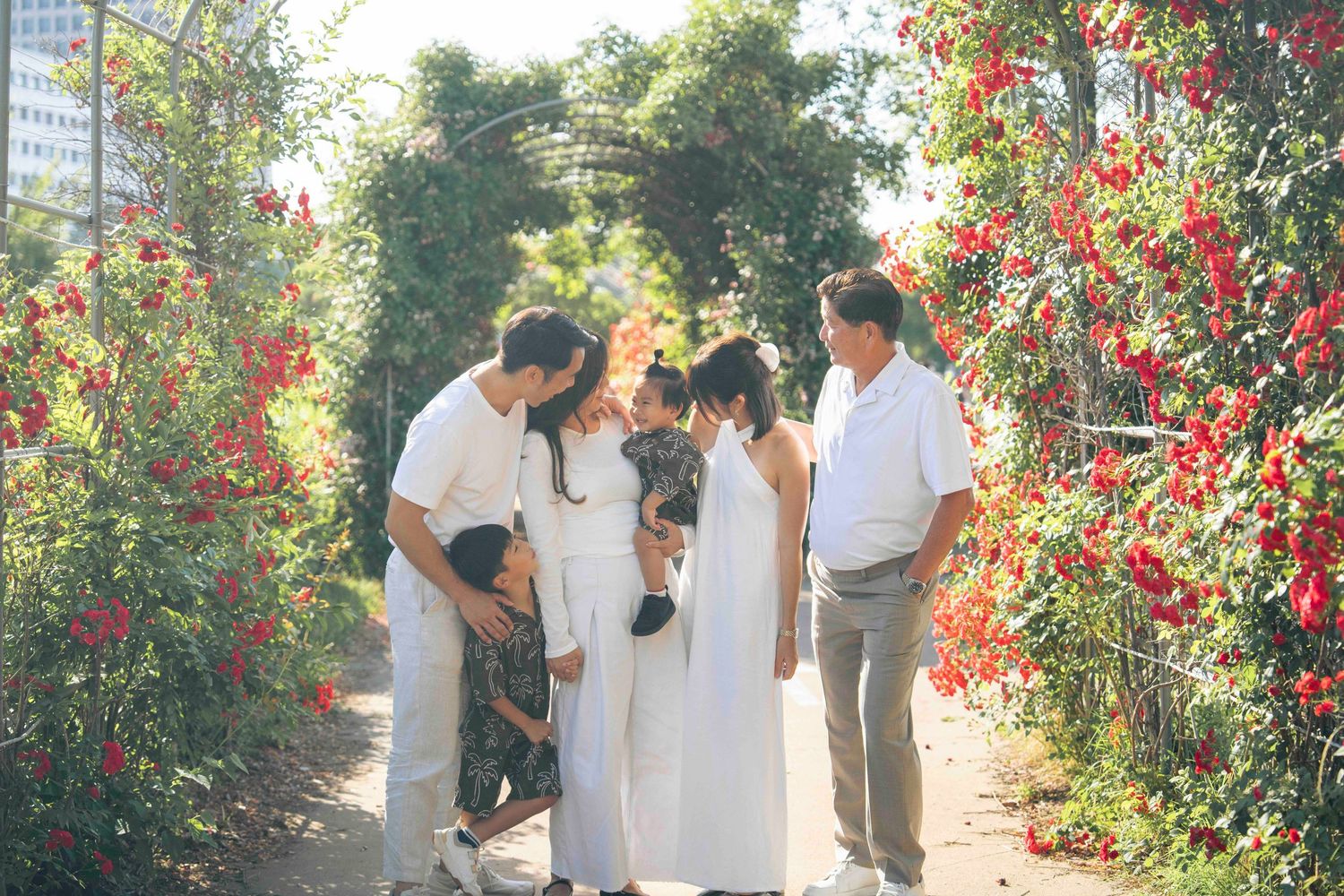 Family gathering in a flower garden with red blooms and green arch, dressed in white and gray outfits.