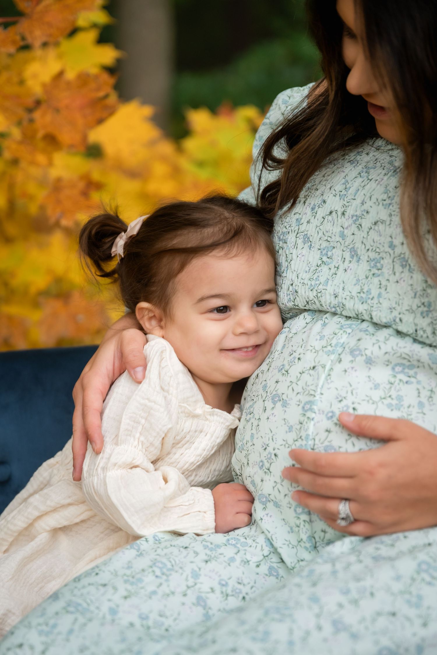 A young girl smiles while hugging her pregnant mother's belly outdoors among vibrant autumn foliage.