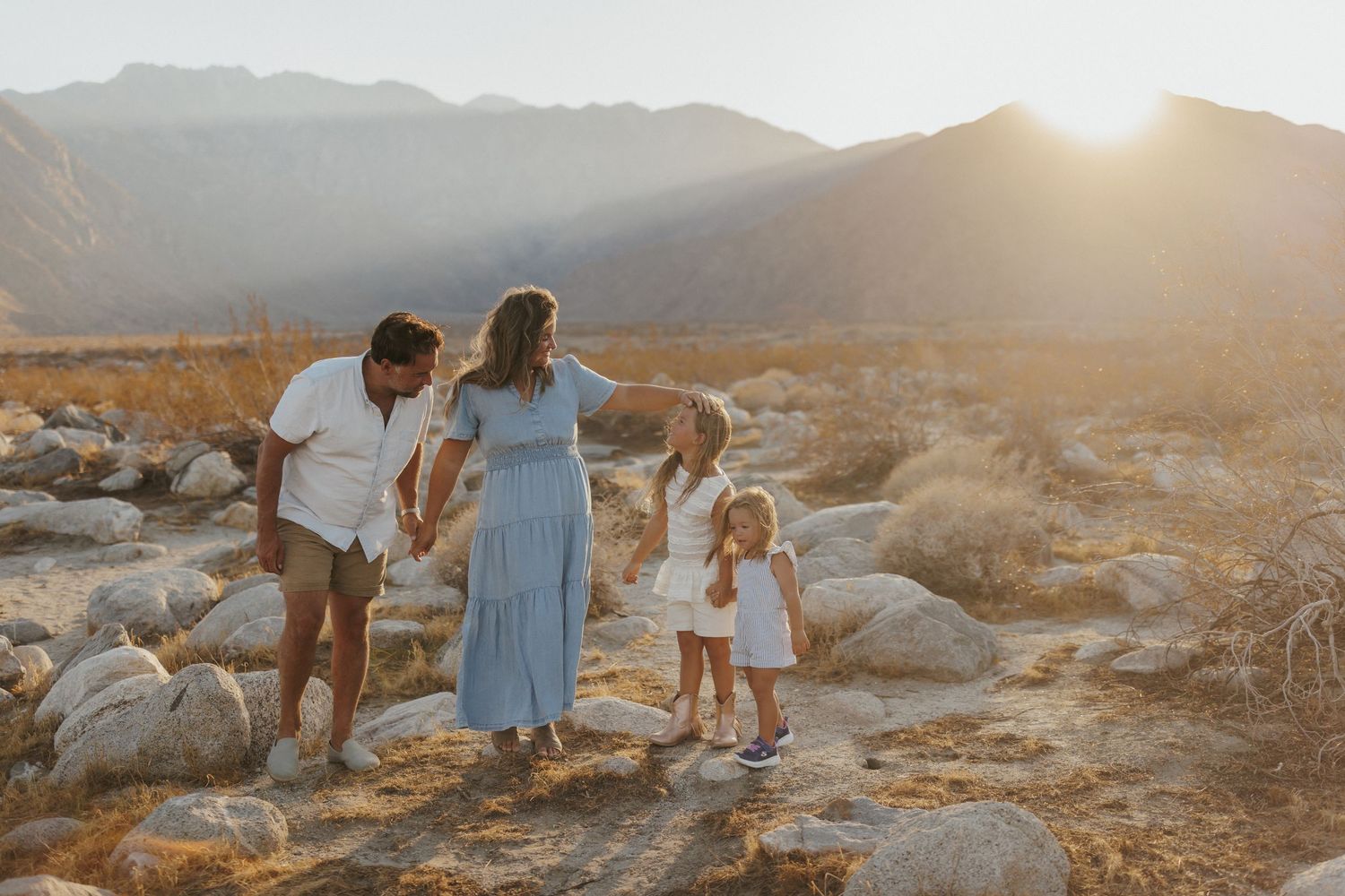 Family of four holding hands at golden hour with mountain backdrop on rocky terrain.