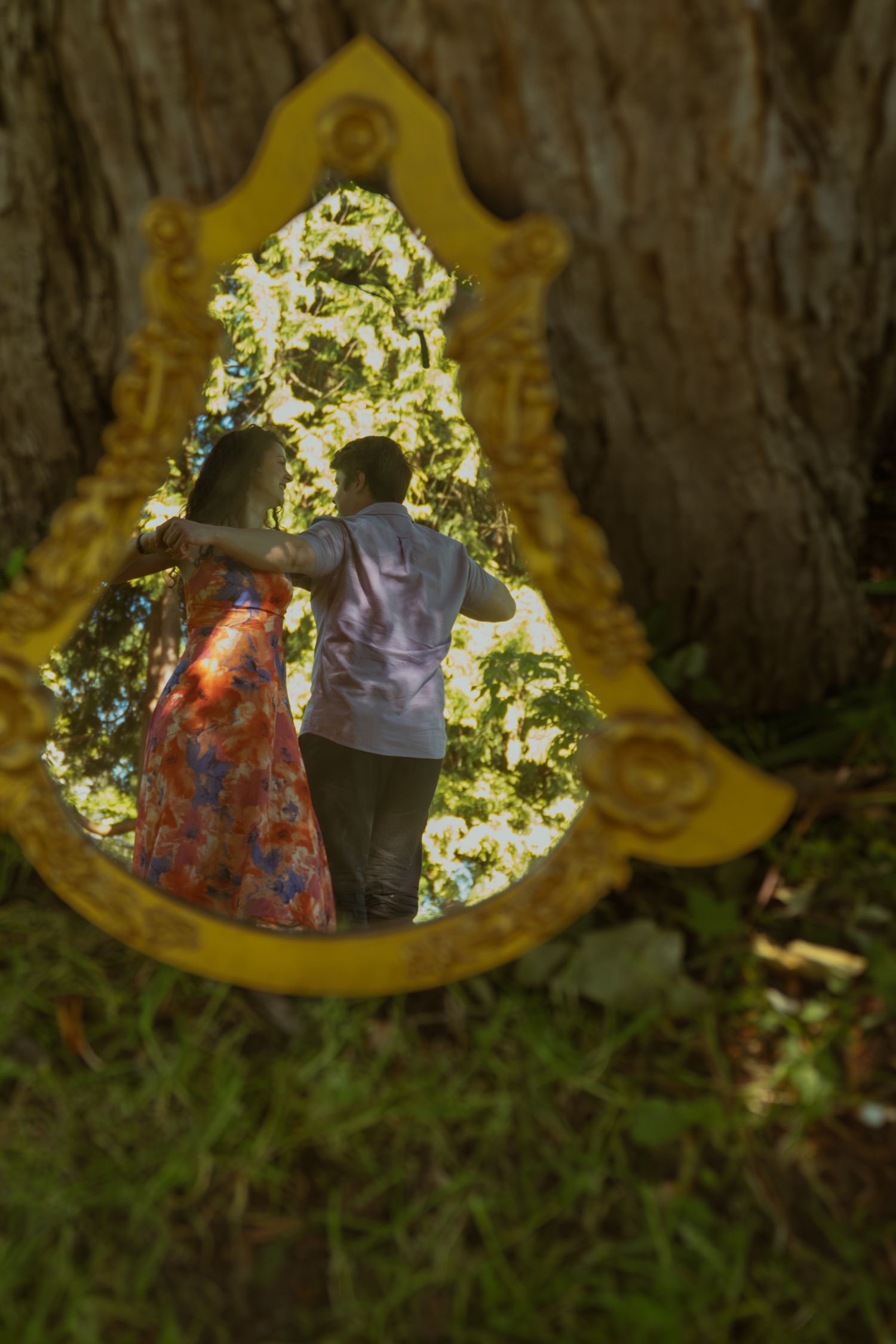 Couple embracing reflected in ornate yellow mirror leaning against tree outdoors.