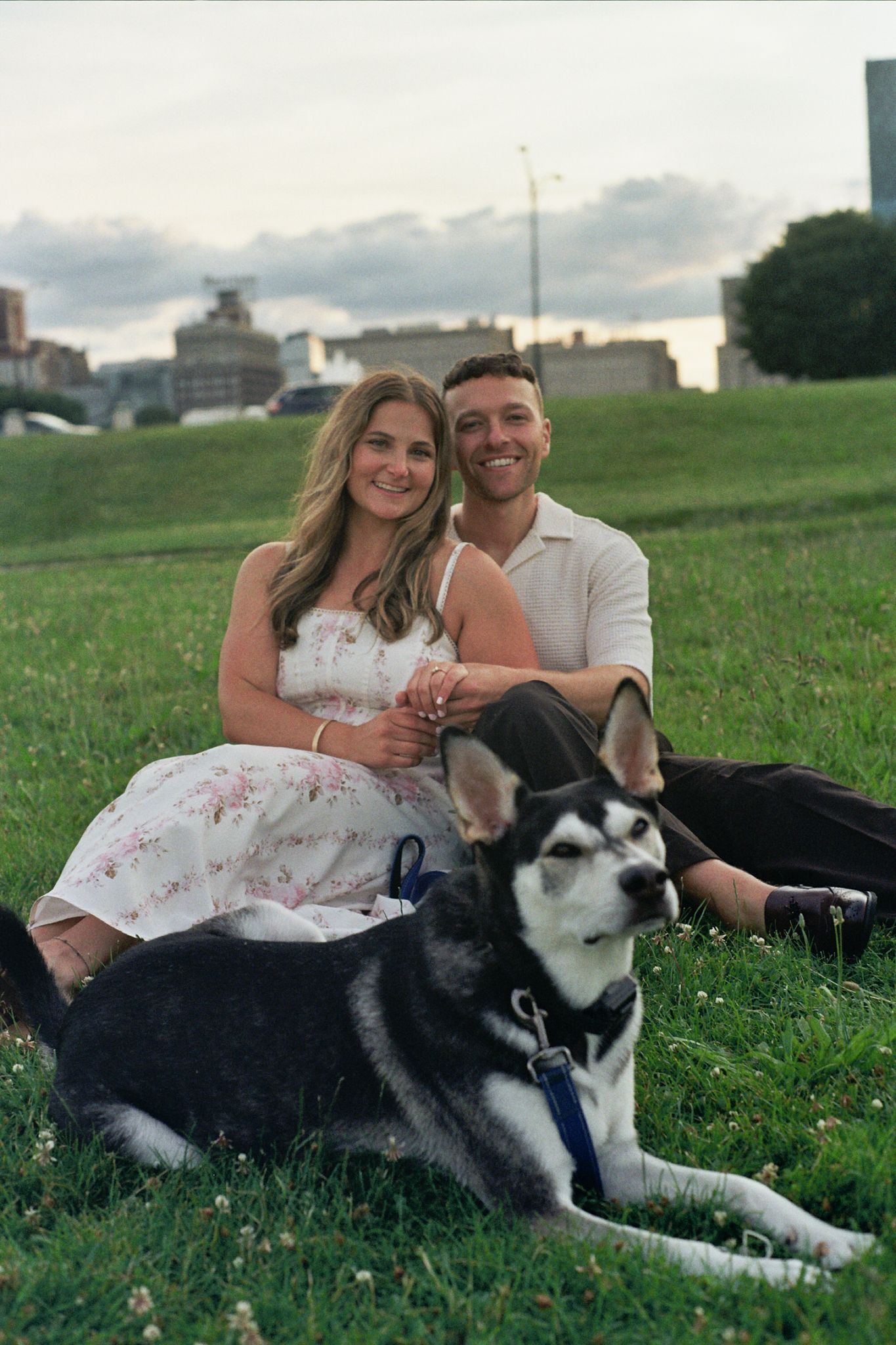 Couple sitting on grass with a Husky dog in an urban park at dusk, woman in floral dress and man in white shirt.