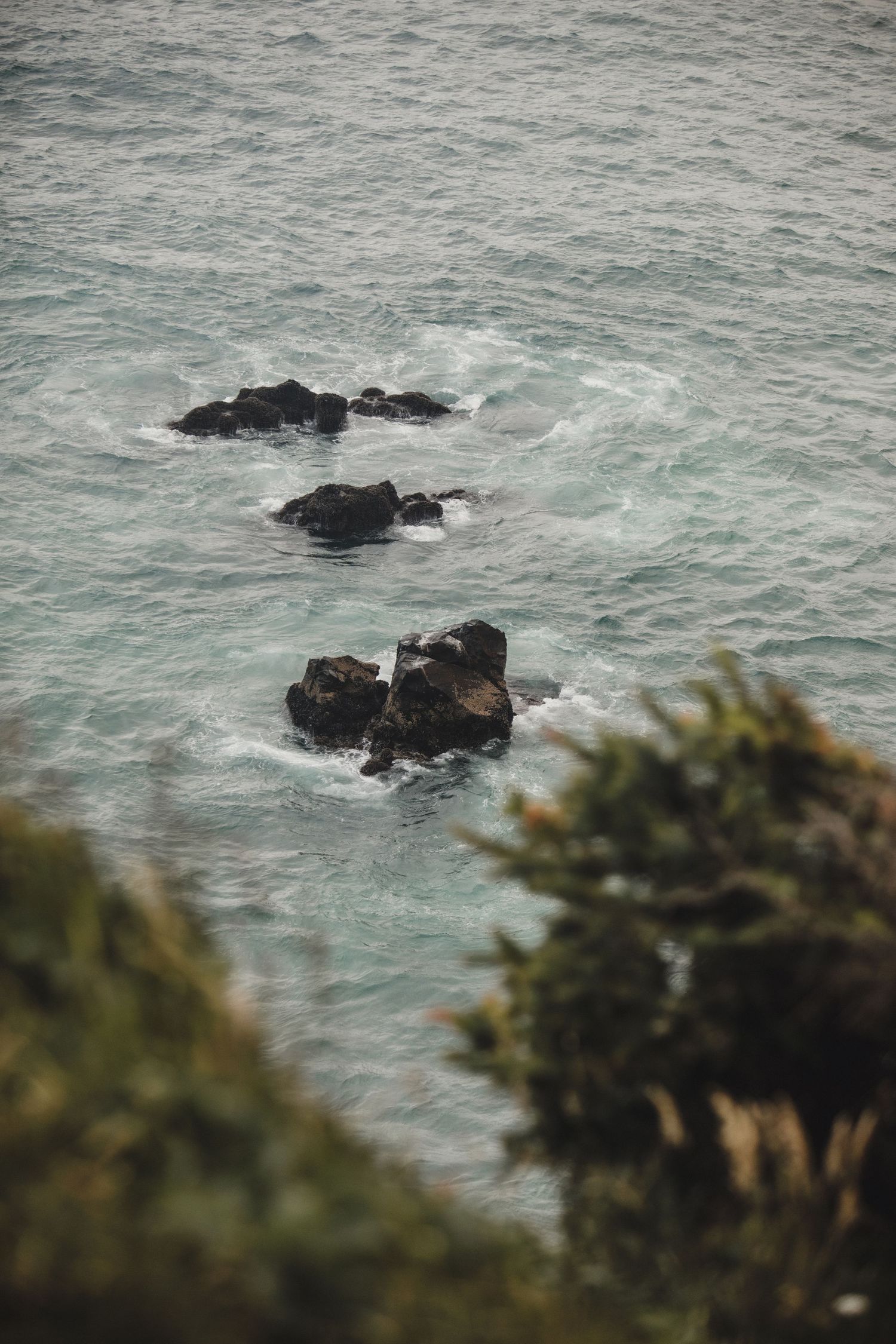 Dramatic coastal cliffs with green tops drop sharply into churning turquoise ocean waves below an overcast sky.