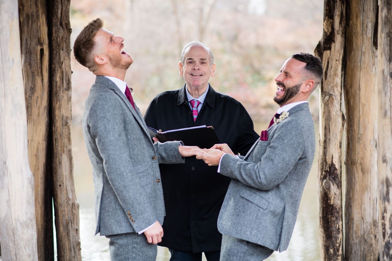 Two grooms in gray suits laugh joyfully during their wedding ceremony with an officiant between them.
