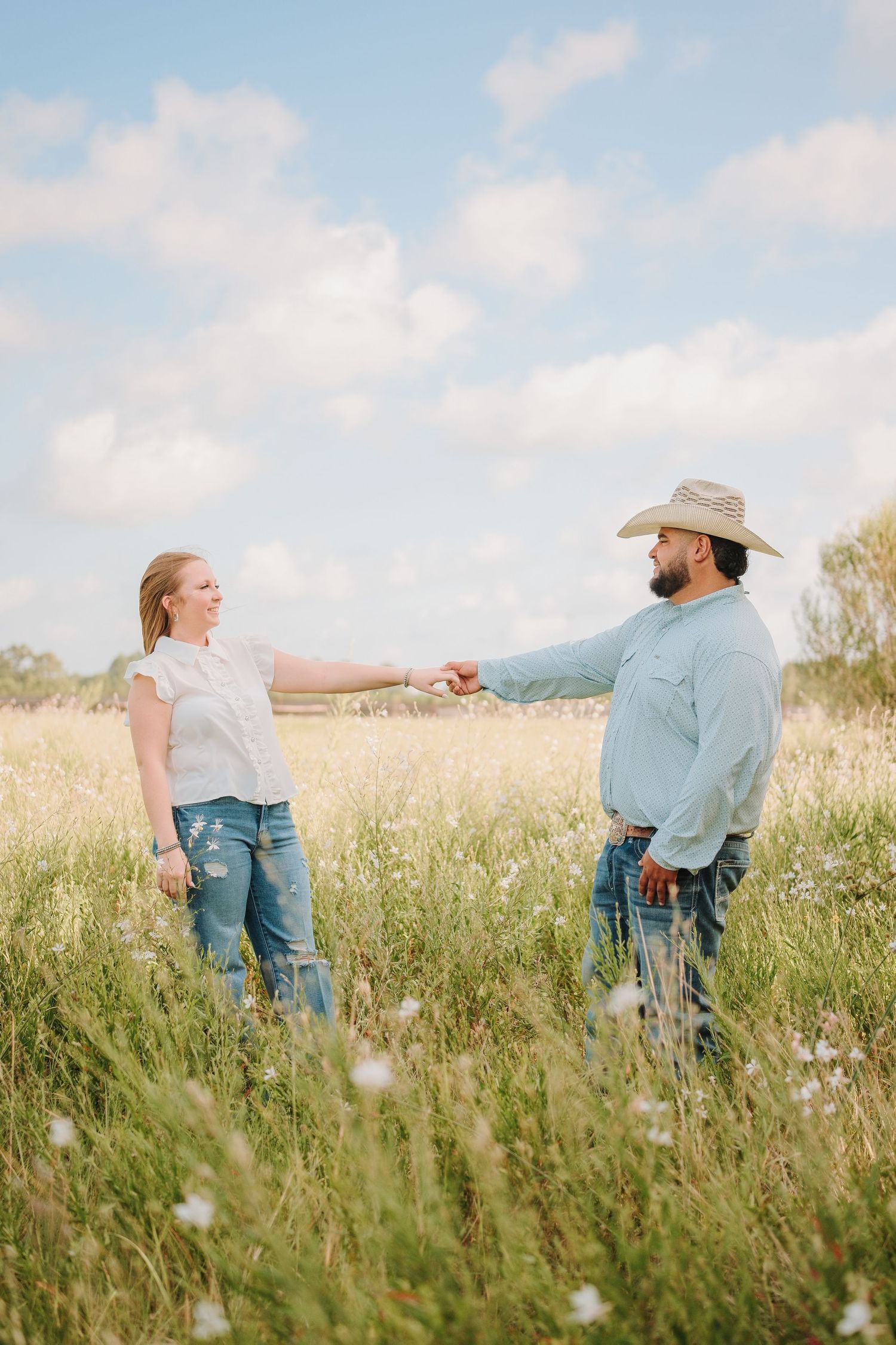 Couple holding hands in a wildflower field, woman in white blouse and jeans, man in cowboy hat and blue shirt.