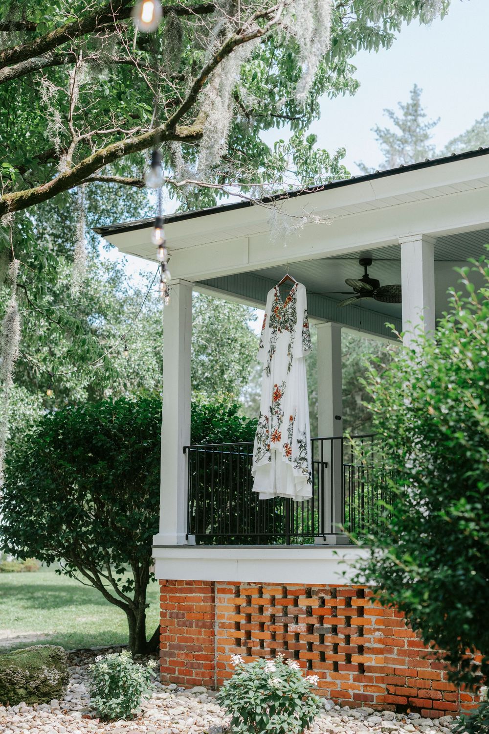 Floral wedding dress hanging on a white porch railing surrounded by lush green trees and brick foundation.