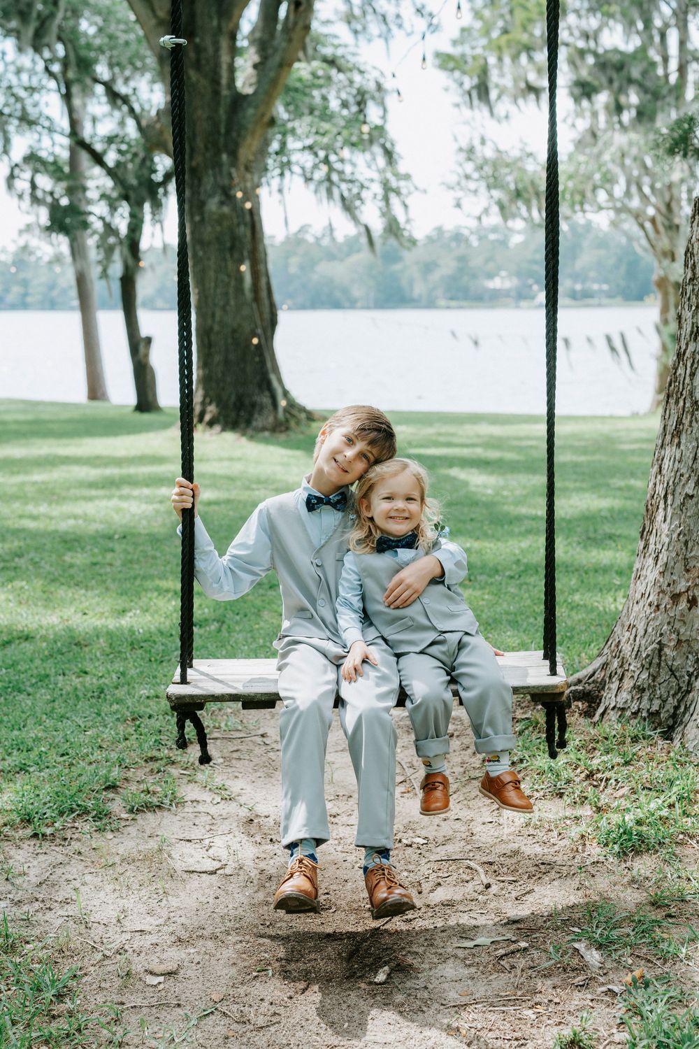 Two boys in matching light blue suits sit together on a wooden garden swing near a serene lakeside setting.