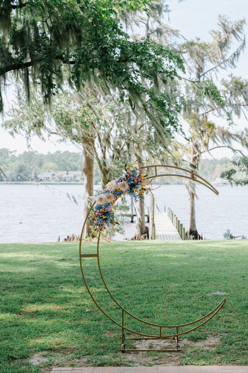 Gold crescent moon wedding arch with colorful flowers by a lakeside, framed by Spanish moss-draped trees.