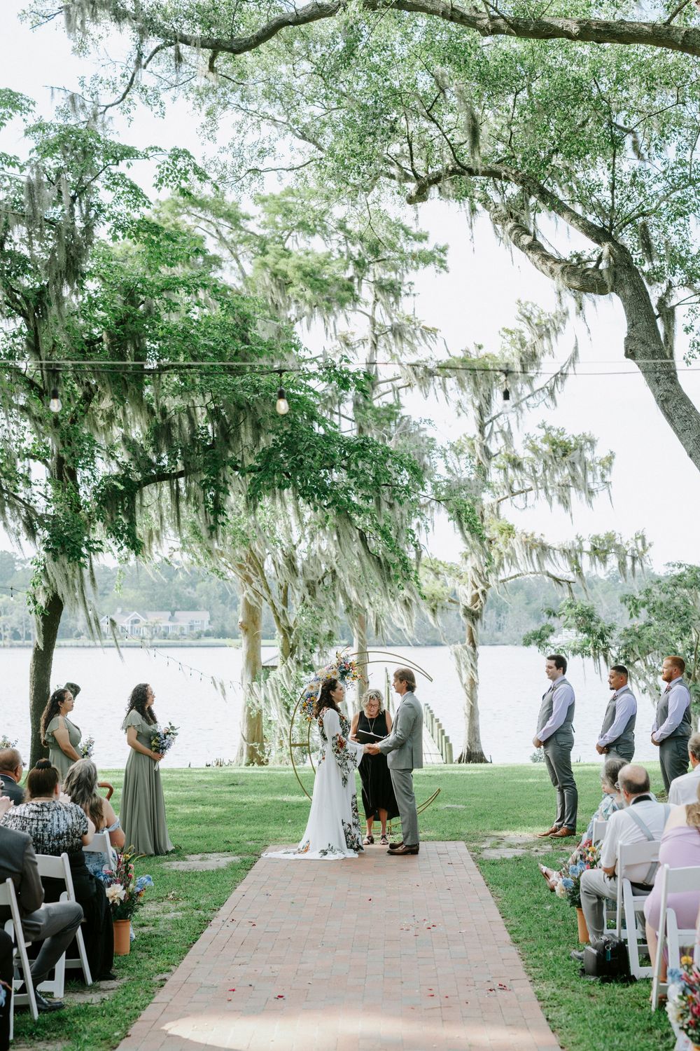Outdoor lakeside wedding ceremony under Spanish moss-draped oak trees with couple at altar on green lawn.