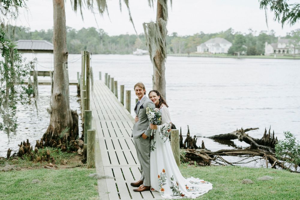 Bride and groom pose on a wooden dock by a serene river, surrounded by lush greenery on their wedding day.