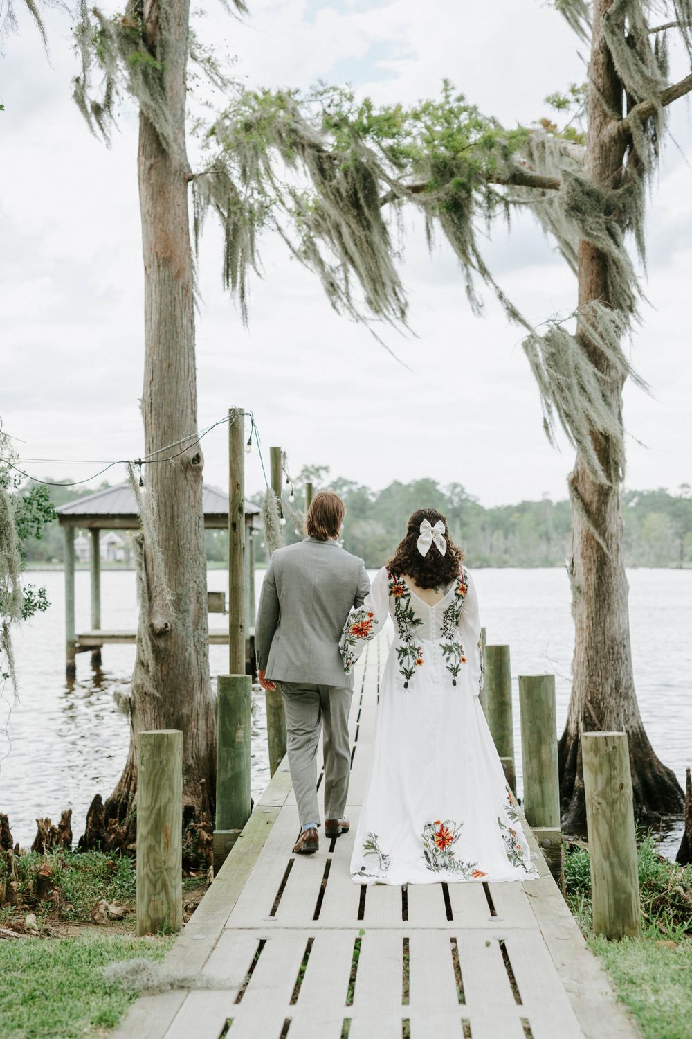 Bride in floral embroidered gown and groom in gray suit walk hand-in-hand down a wooden dock framed by moss-draped cypress trees.