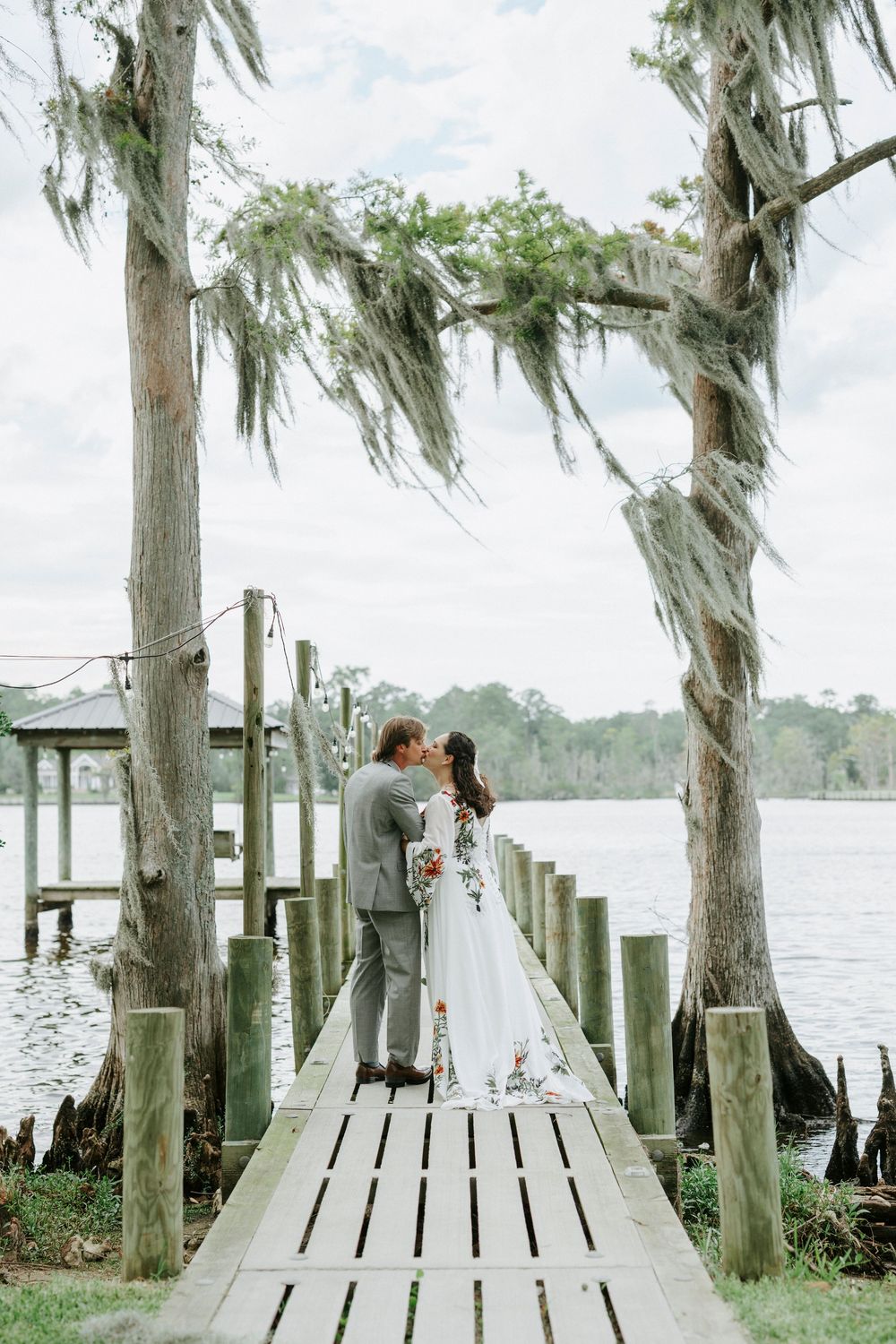 Bride and groom kissing on a wooden dock framed by moss-draped cypress trees beside a tranquil lake.
