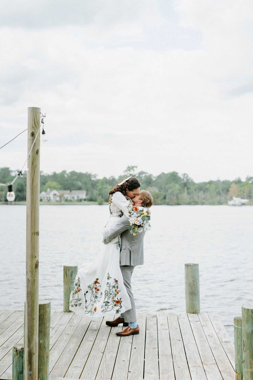 Groom lifting bride in a floral wedding dress on a lakeside dock, sharing a romantic kiss outdoors.