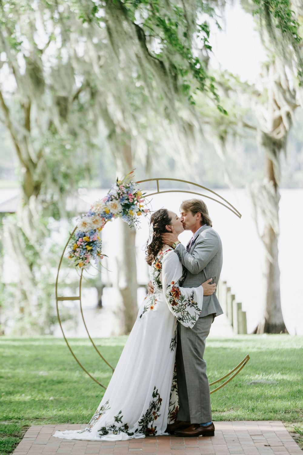 Bride and groom share a kiss under a circular floral arch by a lakeside, bride wearing a floral gown.