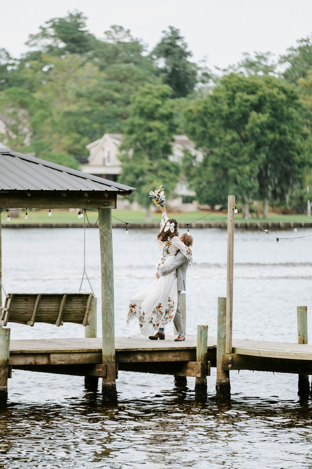 Bride and groom embrace on a waterfront dock, bouquet raised high, with a swing and lush trees in the background.