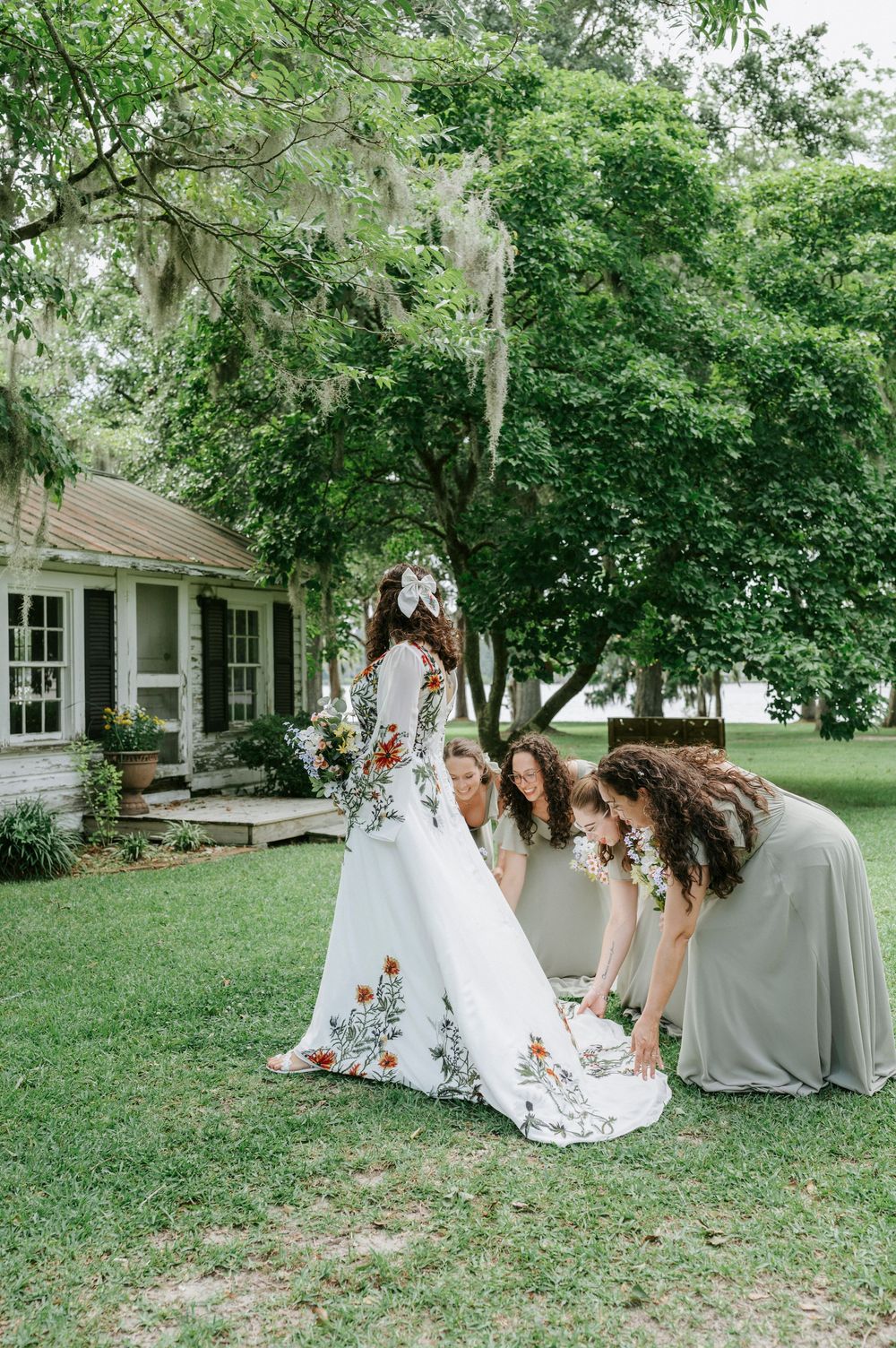 Bridesmaids adjusting floral wedding dress train on green lawn outside rustic Southern home with moss-draped trees.