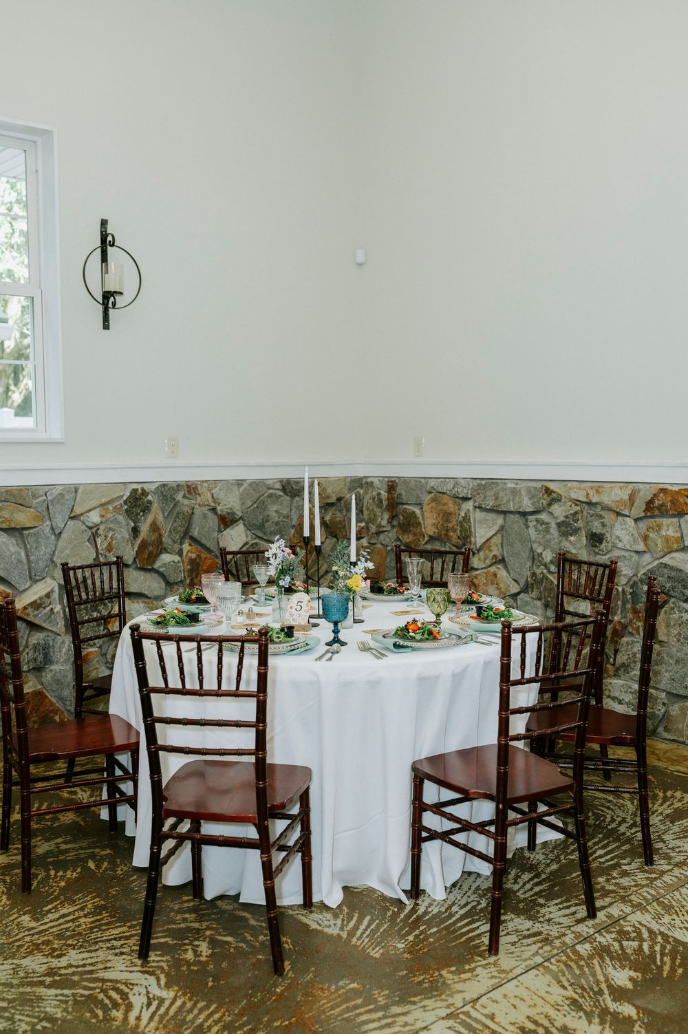 Elegant round dining table with white linen, candles, and floral centerpiece, surrounded by mahogany Chiavari chairs in a stone-walled venue