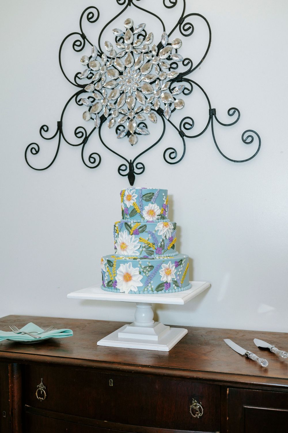 Three-tier floral wedding cake with daisy design on white pedestal stand, displayed on wooden dresser beneath decorative wall art.