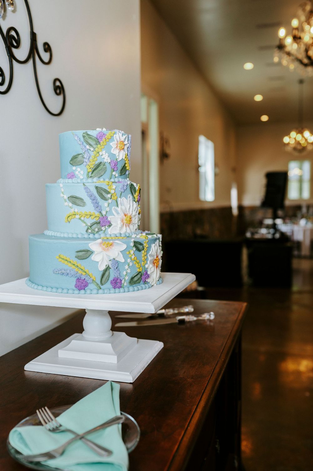 Elegant three-tier light blue wedding cake with hand-painted floral details displayed on a white pedestal stand.
