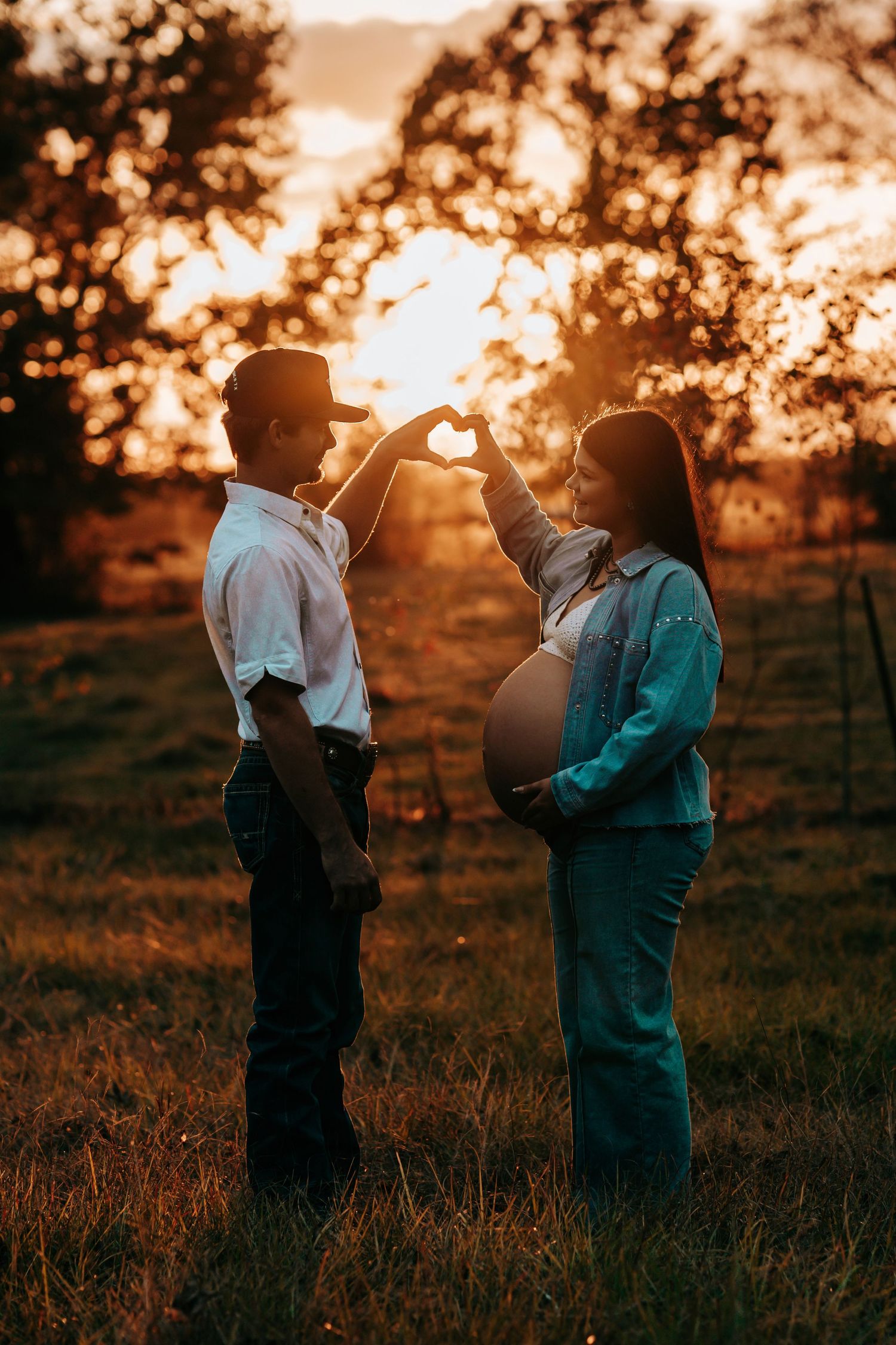 Couple making heart shape with hands at golden sunset, pregnant woman in teal outfit with partner in white shirt in field.
