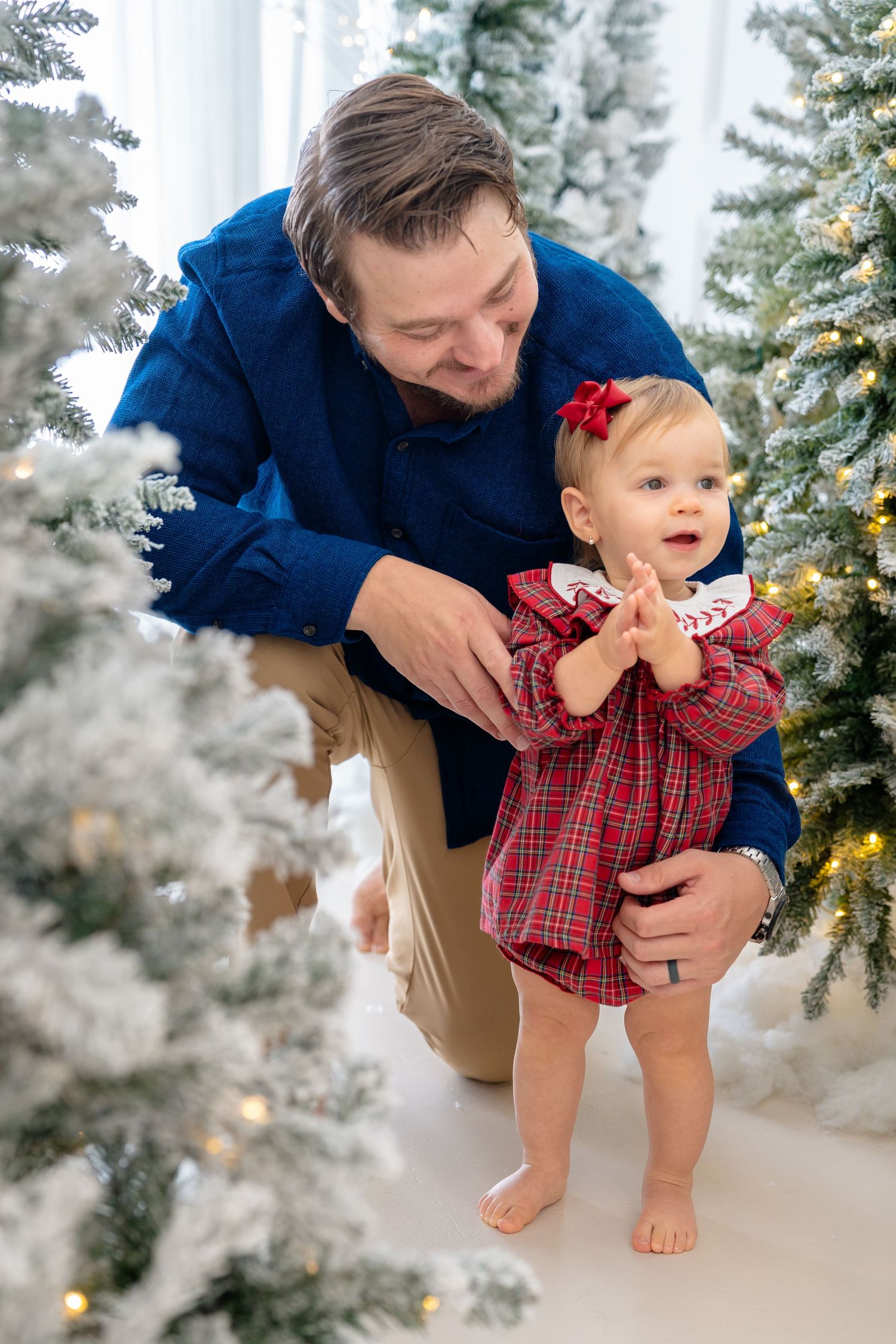 Father helps baby girl in red plaid dress stand near snow-flocked Christmas trees with warm glowing lights.