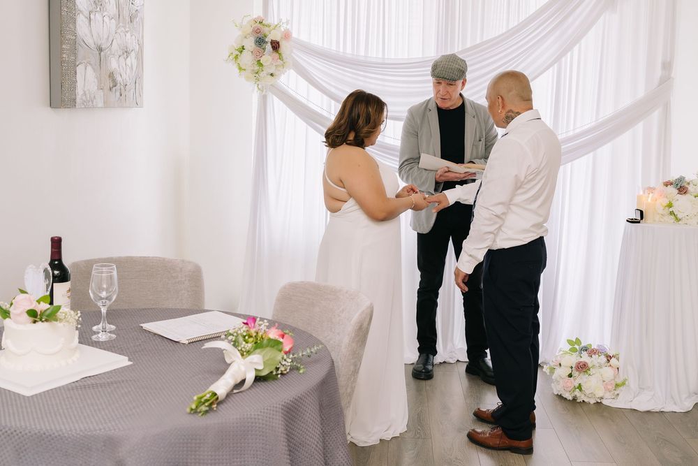 Couple exchanging vows with officiant in elegant white wedding venue decorated with floral arrangements and draped fabric.