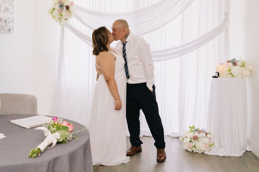 Bride and groom sharing a kiss at wedding ceremony with white draped backdrop and floral arrangements.