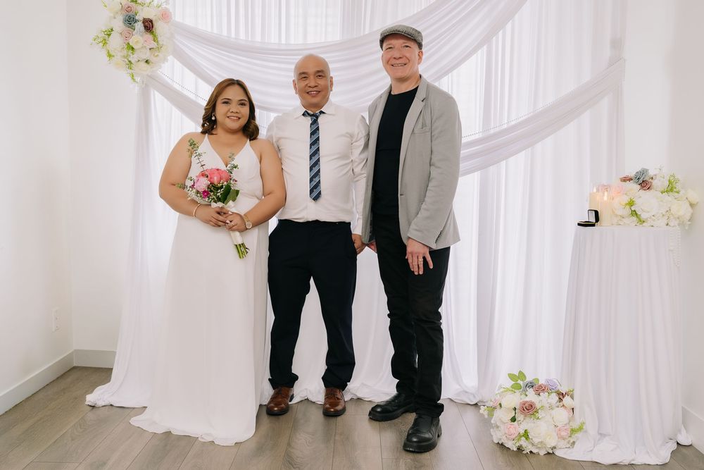 Bride in white gown holding bouquet poses with groom in blue tie and guest at elegant wedding ceremony backdrop.