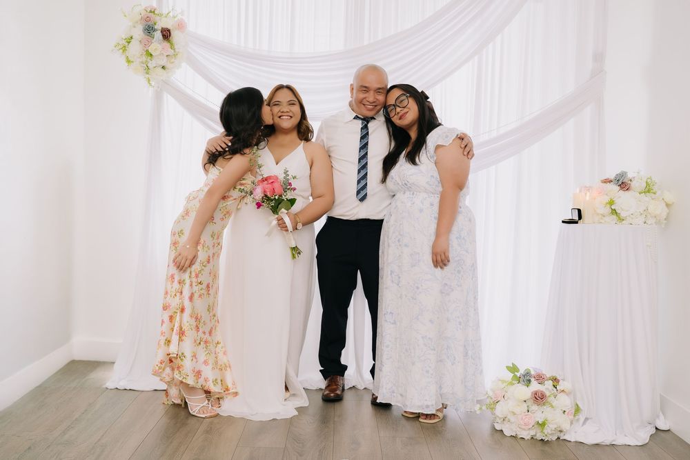 Family celebrating a wedding ceremony, posing joyfully in front of elegant white drape backdrop with floral arrangements.