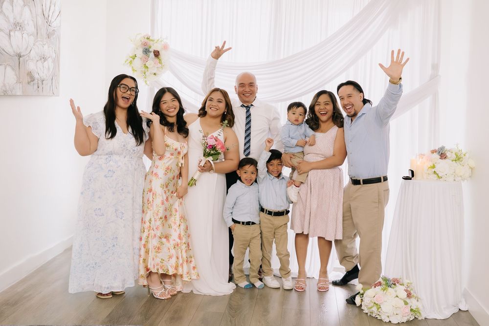 Family celebrating a wedding in a bright white venue, posing joyfully with bride holding bouquet.