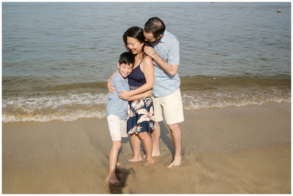 Family of three embracing on a Sayulita beach in Mexico with gentle ocean waves, candid lifestyle family photography Riviera Nayarit.