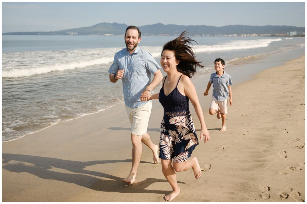 Family of three running on Sayulita beach, Mexico with ocean waves and mountain views, candid Riviera Nayarit family photography.