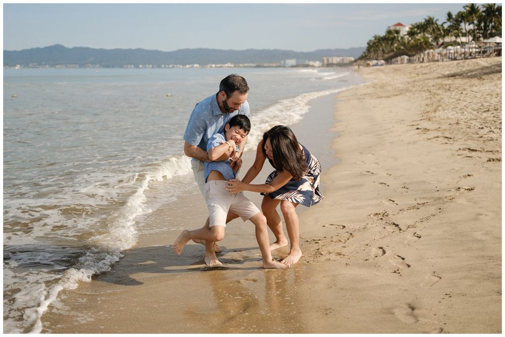 Family of three playing at ocean’s edge in Sayulita, Mexico with waves over feet, candid beach family photography Riviera Nayarit.