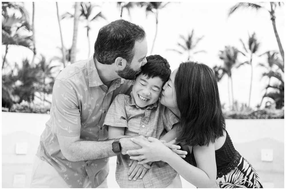 Black and white family portrait in Punta Mita, Mexico: parents kissing laughing son outdoors near palm trees, candid beach photography.