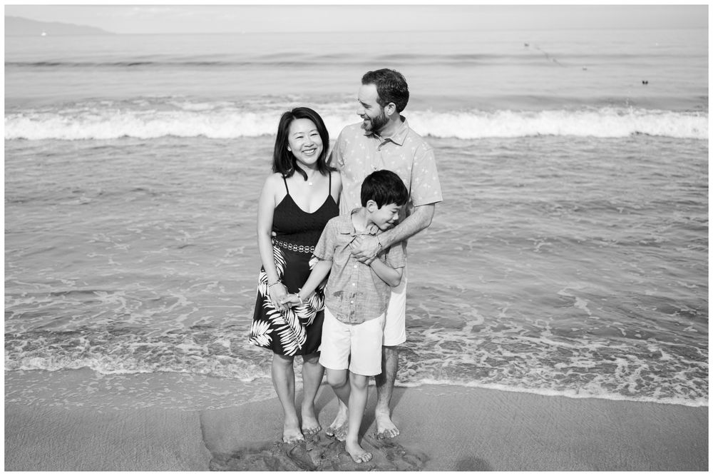 Black and white family of three posing on Sayulita beach, Mexico with ocean waves, Riviera Nayarit family photographer session.