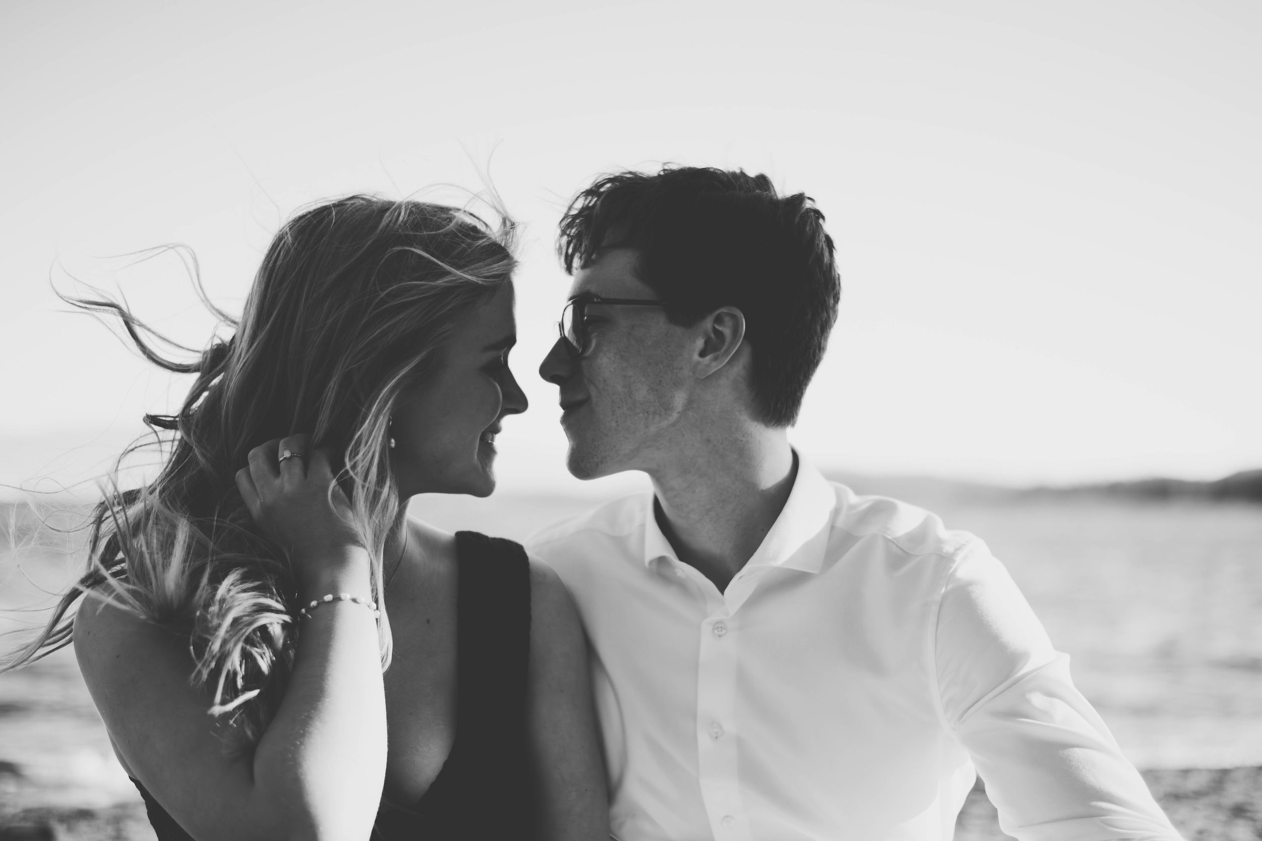 Black and white photo of a romantic couple leaning close together on a beach, with wind blowing through the womans hair.